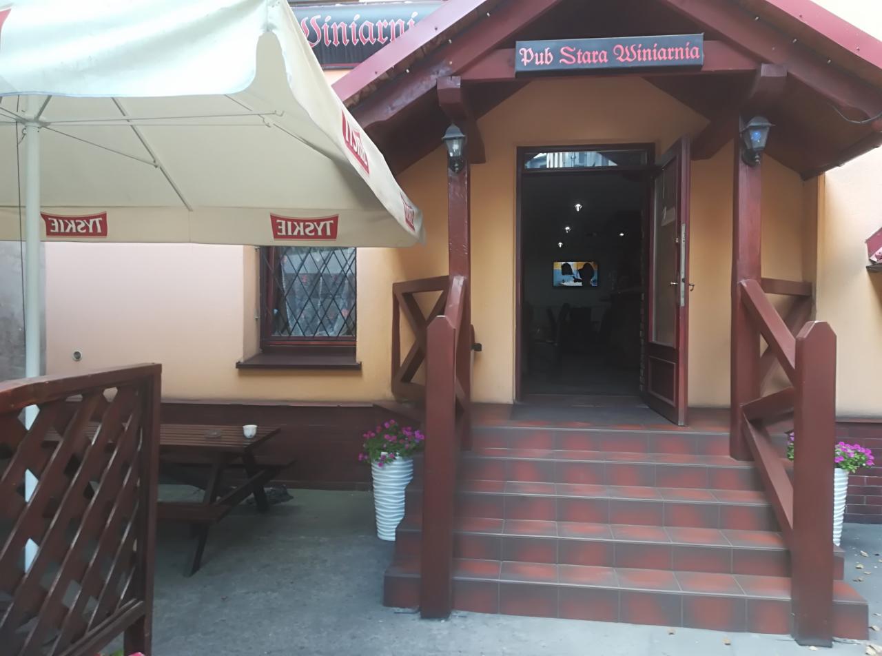 Wejście do pubu Stara Winiarnia z zewnętrznym stolikiem pod parasolem, widoczne schody z płytek i kwiaty w donicach.