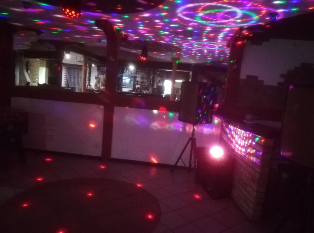Wnętrze lokalu z barem, oświetlone kolorowymi światłami dyskotekowymi, głośnik na statywie, ceglana dekoracja baru.
