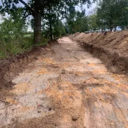 Wykopany pas terenu wzdłuż drzew, przygotowany pod budowę lub utwardzenie drogi gruntowej, z widocznymi śladami po maszynach i pryzmami ziemi po bokach.