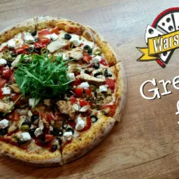 Pizza grecka fit z kurczakiem, fetą, oliwkami, pomidorami i rukolą na drewnianym stole z logotypem firmy w tle.