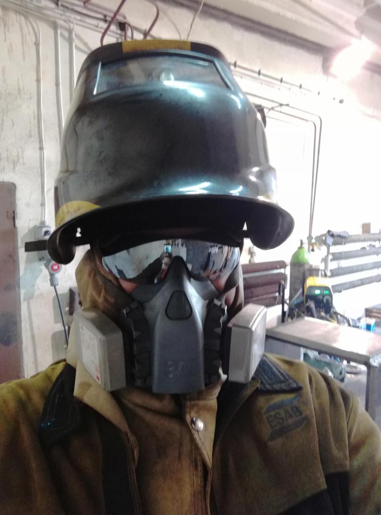 Osoba w pełnym sprzęcie ochronnym do spawania: maska z filtrami, gogle odblaskowe i hełm spawalniczy, widoczna kurtka robocza ESAB, w tle stanowisko pracy ze spawarką.