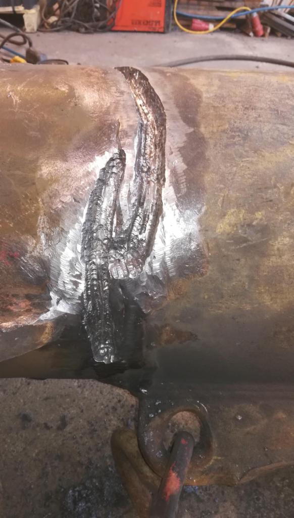 Świeża spoina na zardzewiałej, grubej rurze, widoczne ślady obróbki i uchwyt mocujący.