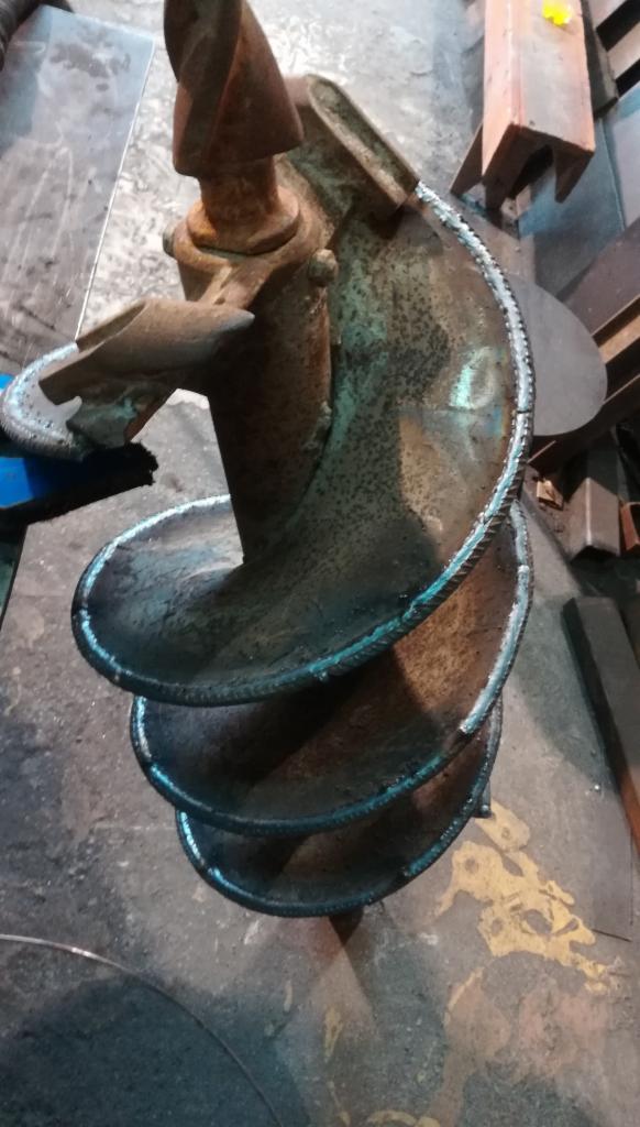 Stara, zardzewiała wiertnica ślimakowa z widocznymi śladami spawania na krawędziach spirali, leżąca na brudnej, betonowej podłodze.