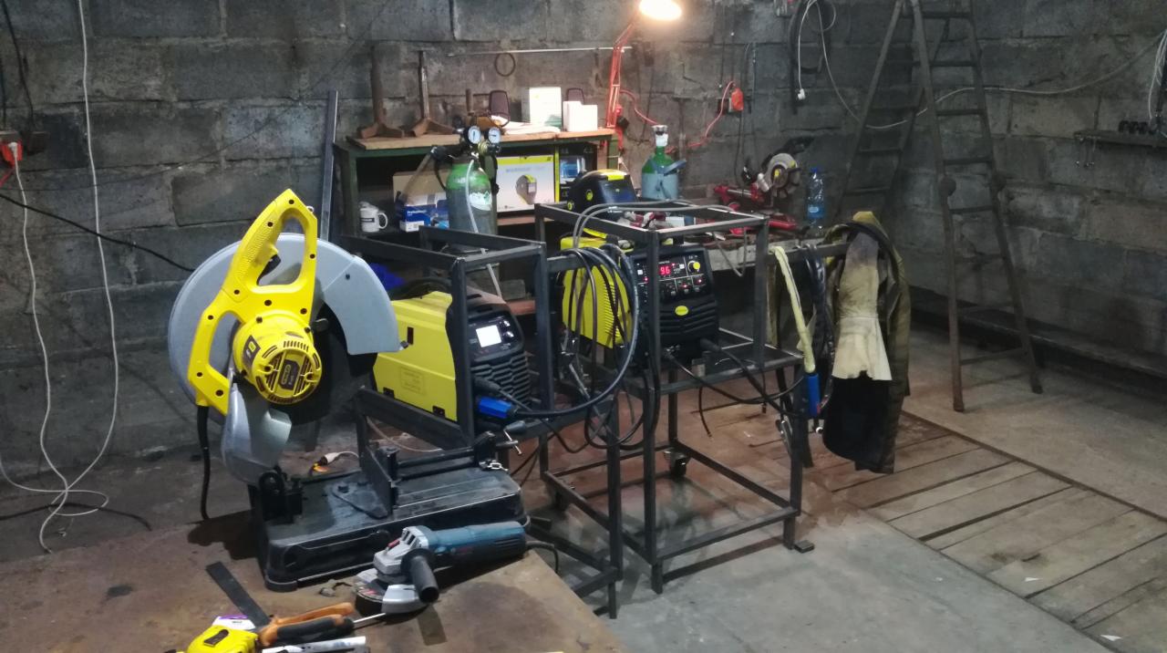 Warsztat z żółtą przecinarką do metalu, szlifierką kątową i spawarkami na metalowych wózkach na tle murowanej ściany i drabiny.