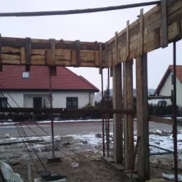 Szare bloczki betonowe z widocznym zbrojeniem i drewnianym szalunkiem na budowie domu jednorodzinnego, w tle ośnieżona ulica i domy z czerwonymi dachami.