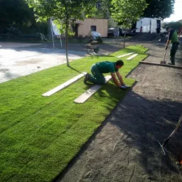 Układanie trawy z rolki na przygotowanym terenie przy placu zabaw, widoczni dwaj pracownicy w zielonych uniformach.
