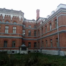 Ceglany budynek zabytkowy z wieżyczkami i wysokim kominem, widoczny z poziomu gruntu, z fragmentem zarośniętego terenu przed nim.