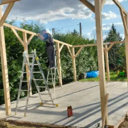 Konstrukcja drewniana altany w trakcie montażu, pracownik na aluminiowej drabinie przytwierdza elementy, zielone tuje w tle.