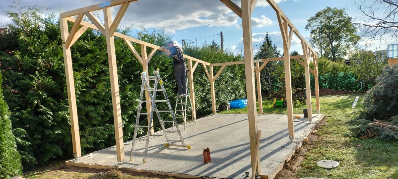 Konstrukcja drewniana altany w trakcie montażu, pracownik na aluminiowej drabinie przytwierdza elementy, zielone tuje w tle.