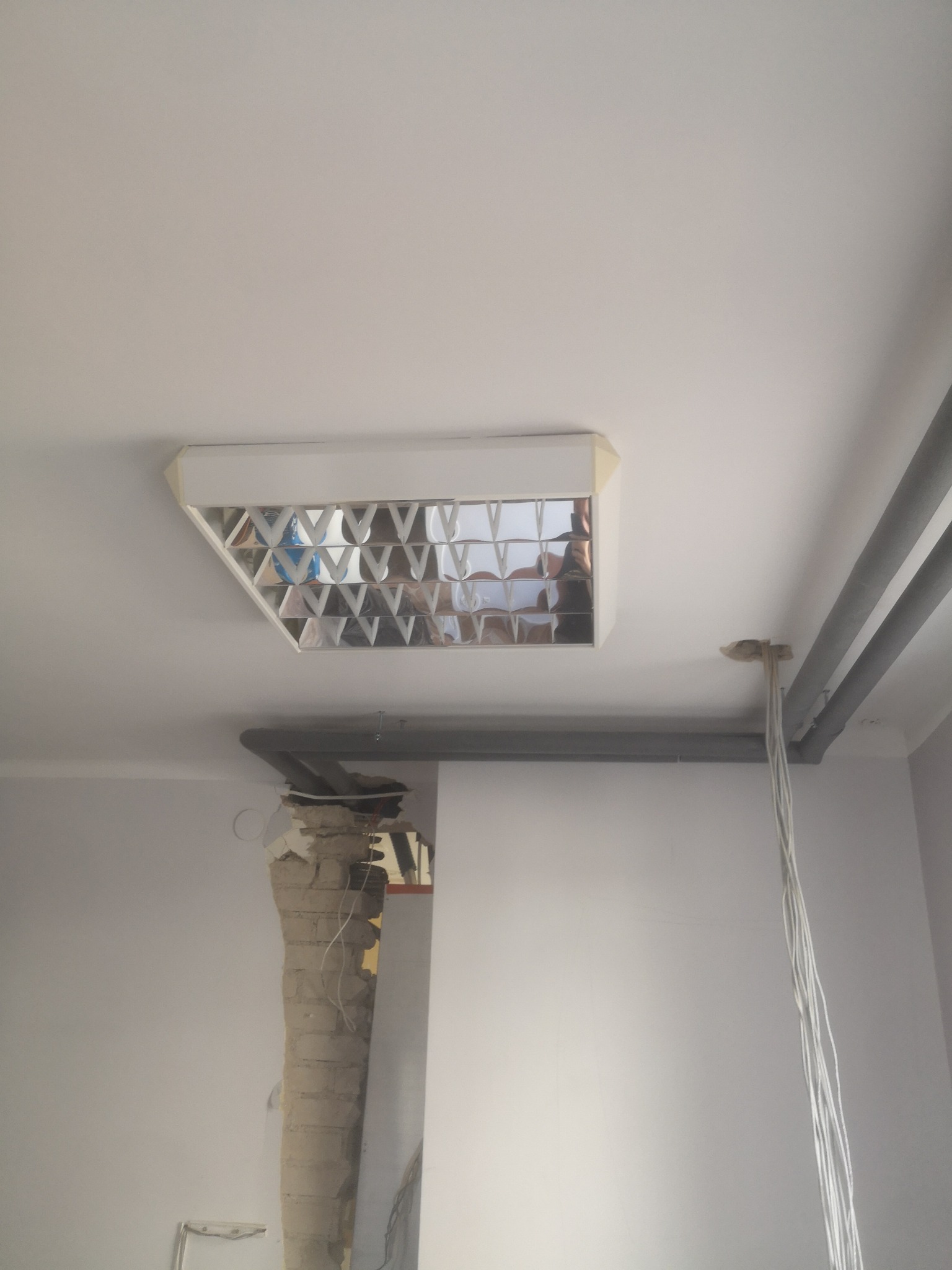 Widok sufitu w trakcie remontu z odsłoniętymi rurami, przewodami i uszkodzonym fragmentem ściany, widoczna lampa rastrowa z odbiciem osoby.