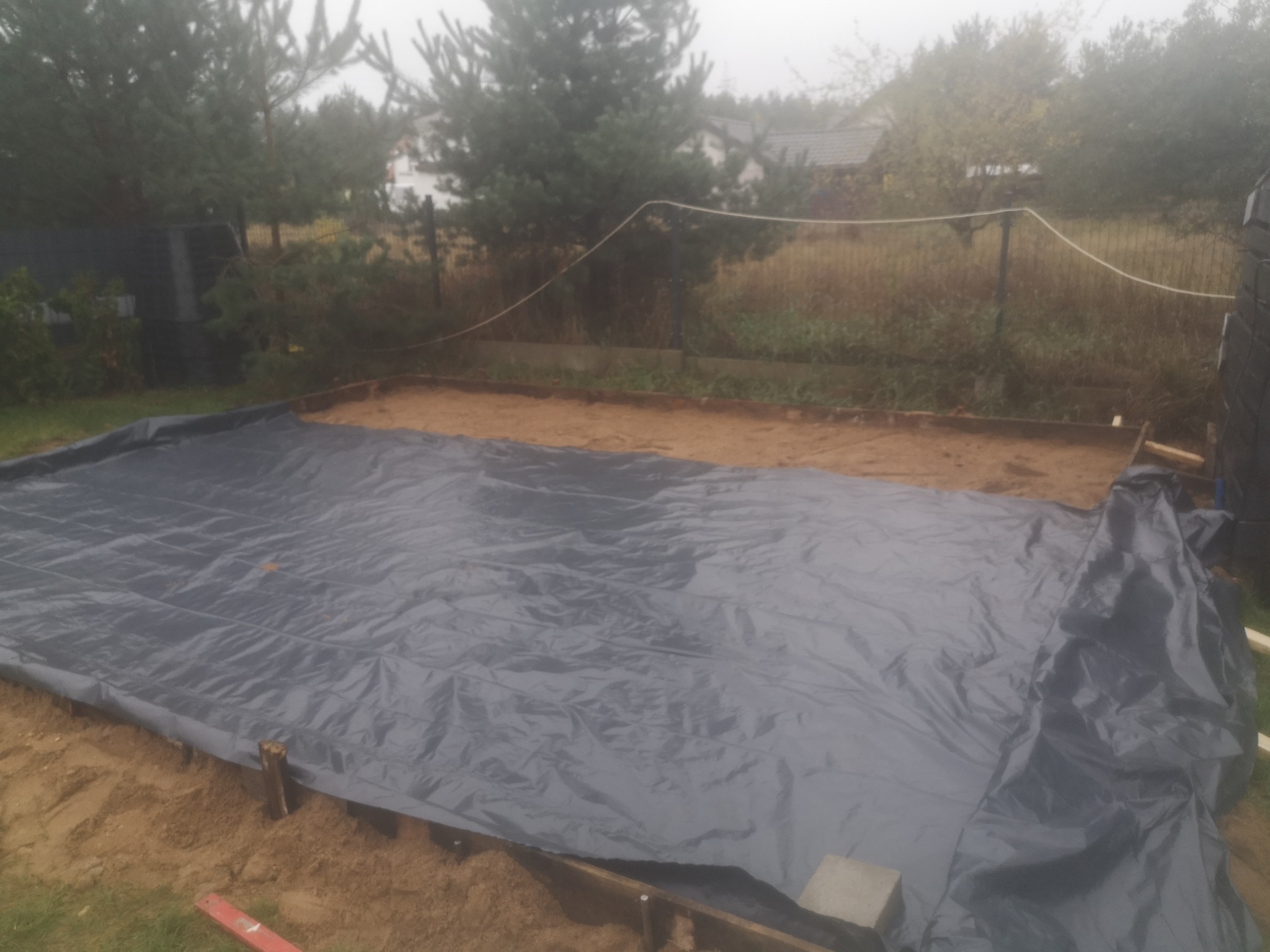 Przygotowany wykop z drewnianą szalunką i rozłożoną folią ochronną, widok na zewnątrz w pochmurny dzień.