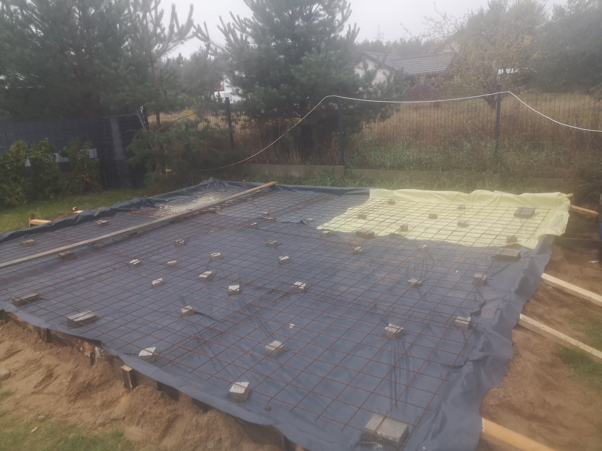 Przygotowanie terenu pod wylewkę fundamentów: siatka zbrojeniowa na folii, podkłady z kostki betonowej, drewniane szalunki, widok z góry w pochmurny dzień.