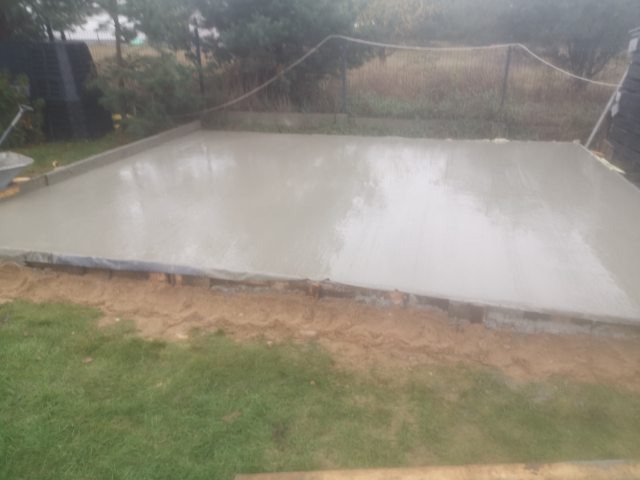 Świeżo wylana, mokra płyta betonowa na przygotowanym podłożu z widocznym szalunkiem, otoczona trawnikiem i elementami krajobrazu.