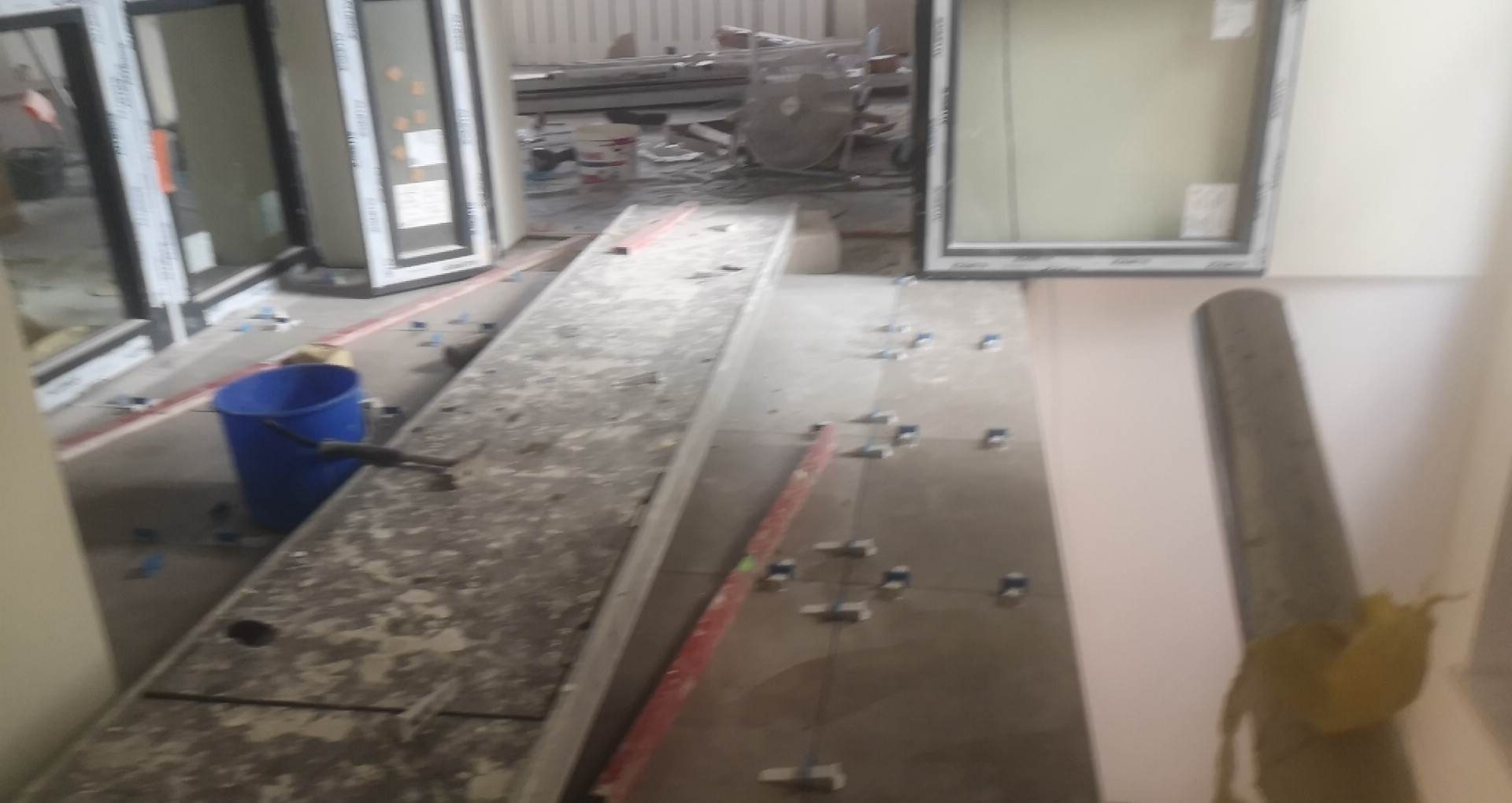 Przygotowanie do układania płytek podłogowych: poziomowanie powierzchni, niebieskie wiadro z narzędziami w tle, widoczne krzyżyki dystansowe, poziomica w użyciu.