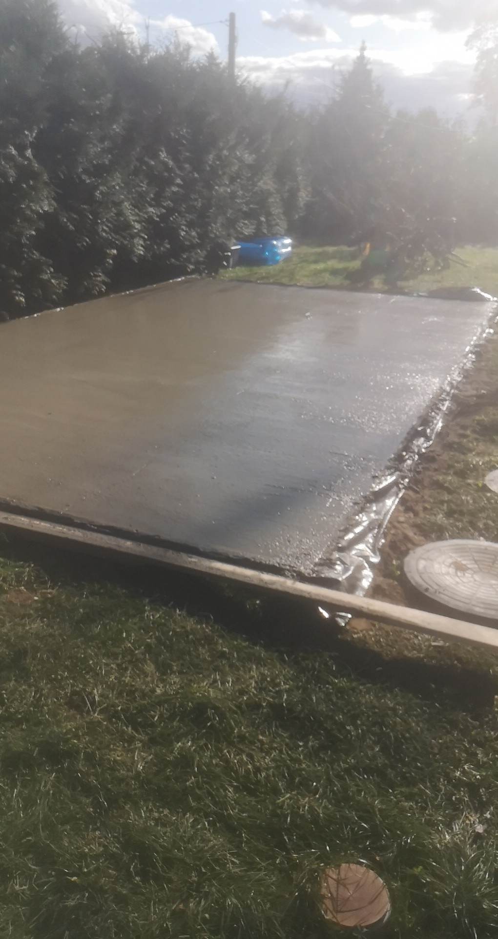 Świeżo wylana, mokra betonowa płyta na zewnątrz, z widocznym szalunkiem i fragmentem trawnika. W tle krzewy i niebieski basen ogrodowy.