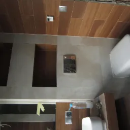 Wnętrze łazienki w trakcie prac wykończeniowych: widoczne płytki imitujące drewno na suficie, częściowo wykończone ściany z wnękami, podwieszana toaleta, lustro z taśmą malarską, butelka...