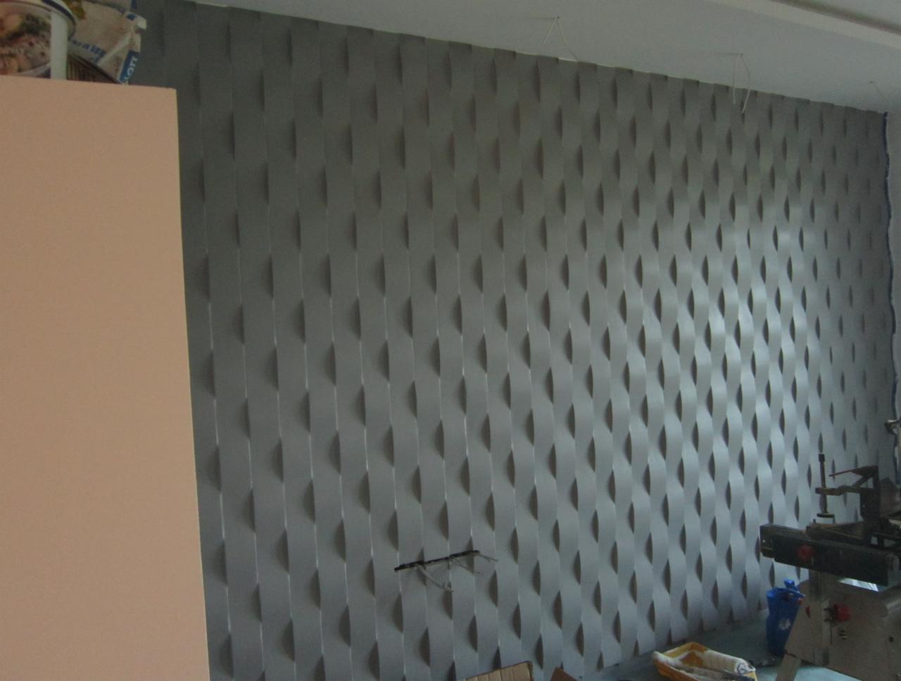 Szara ściana z dekoracyjnymi, trójwymiarowymi panelami ułożonymi w pionowe rzędy, widoczne narzędzia i materiały budowlane w tle.