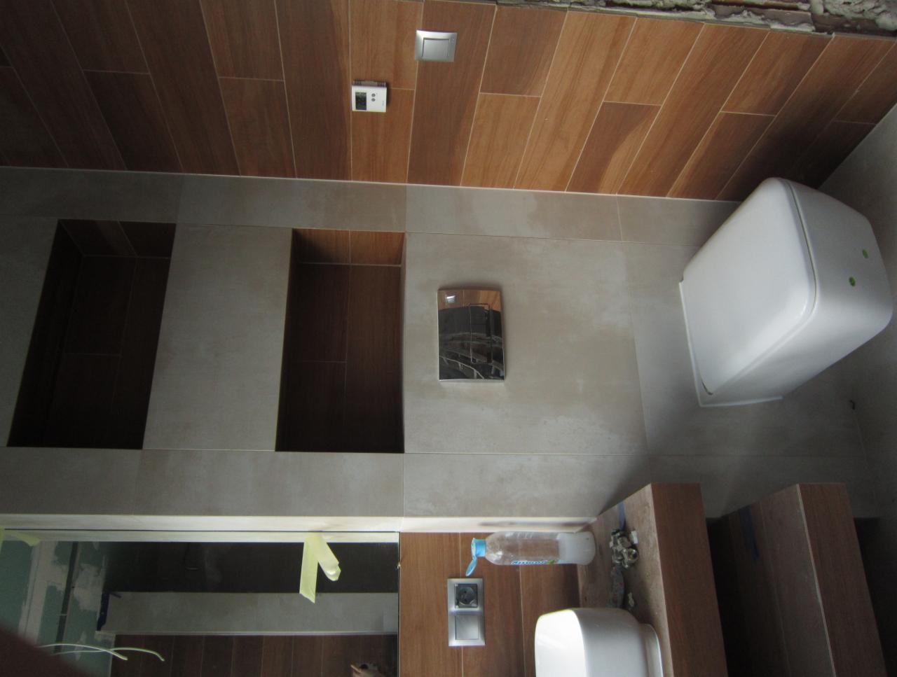 Wnętrze łazienki w trakcie prac wykończeniowych: widoczne płytki imitujące drewno na suficie, częściowo wykończone ściany z wnękami, podwieszana toaleta, lustro z taśmą malarską, butelka...