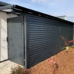 Nowoczesna, grafitowa brama wjazdowa z poziomymi panelami, częściowo otwarta, przylegająca do budynku z zadaszeniem, widok z boku na tle mulczowanej rabaty.