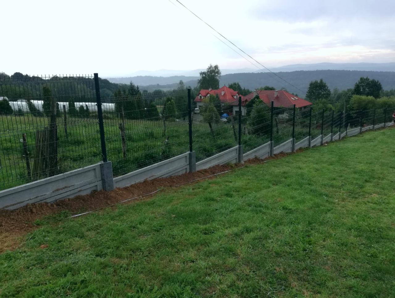 Nowo postawione ogrodzenie z paneli siatkowych na betonowej podmurówce, widok na rozległy krajobraz z domami i wzgórzami w tle, świeżo obsypana ziemia wzdłuż fundamentów.