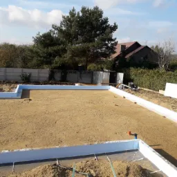 Przygotowany teren pod budowę: wyrównany piasek w obrysie białych płyt izolacyjnych, widoczne elementy instalacji i narzędzia pomiarowe, w tle dom i drzewa.