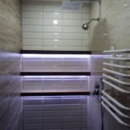 Nowoczesna kabina prysznicowa z białymi, podłużnymi płytkami, podświetlonymi półkami i grzejnikiem drabinkowym.