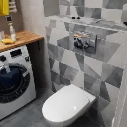 Nowoczesna łazienka z geometrycznymi płytkami w odcieniach szarości, wisząca toaleta, pralka z drewnianym blatem i dozownikiem mydła.