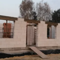 Wznoszenie ścian domu z białych bloczków, widoczne drewniane nadproża okienne i drzwiowe, w tle drzewa i zadaszona altana.