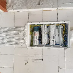 Rozdzielnica elektryczna z zabezpieczeniami, zamontowana na ścianie z bloczków, z widocznymi przewodami w peszlach prowadzonymi wzdłuż ściany, zabezpieczonymi uchwytami.