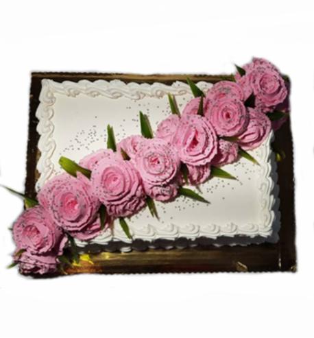 Prostokątny tort z białym lukrem, zdobiony różowymi, cukrowymi kwiatami przypominającymi róże i zielonymi listkami, posypany drobnym makiem.