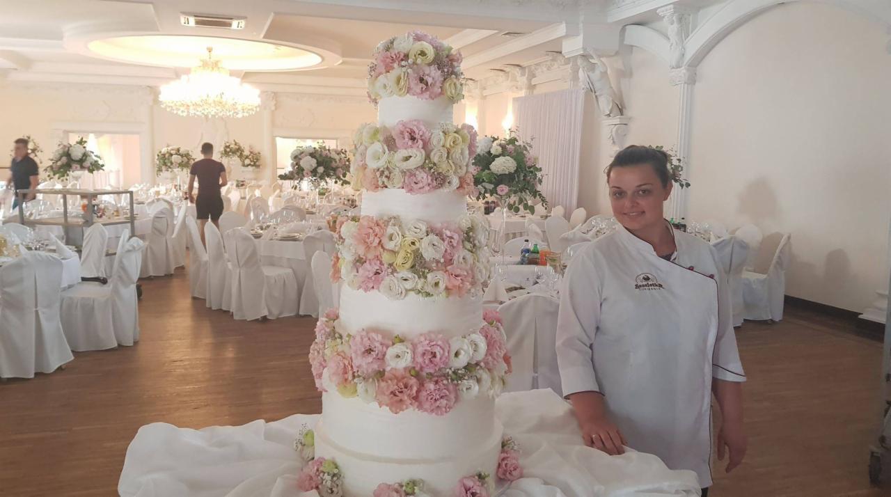 Wielopiętrowy tort weselny zdobiony świeżymi kwiatami w odcieniach różu i bieli, prezentowany przez uśmiechniętą cukierniczkę w fartuchu firmowym na tle sali bankietowej z przygotowanymi stołami.