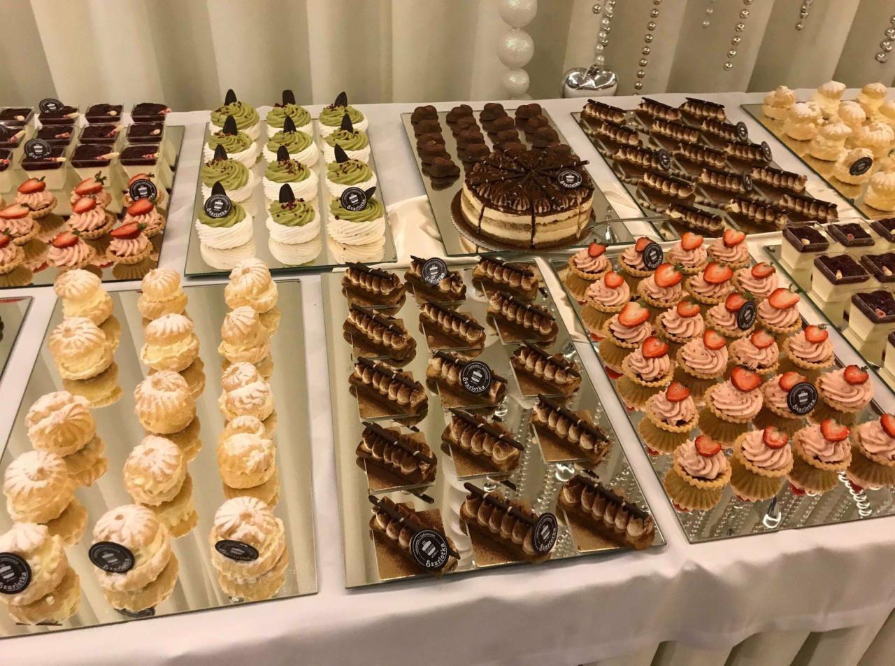 Bogaty wybór deserów na białym stole: eklery z pudrem, bezy z zielonym kremem, mini tartaletki z truskawkami, tiramisu i czekoladowe trufle.