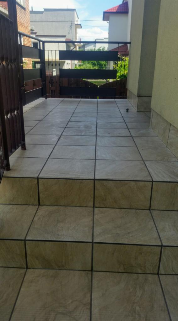 Wyłożony płytkami taras z trzema stopniami, zakończony metalową barierką i drewnianym płotem w tle.