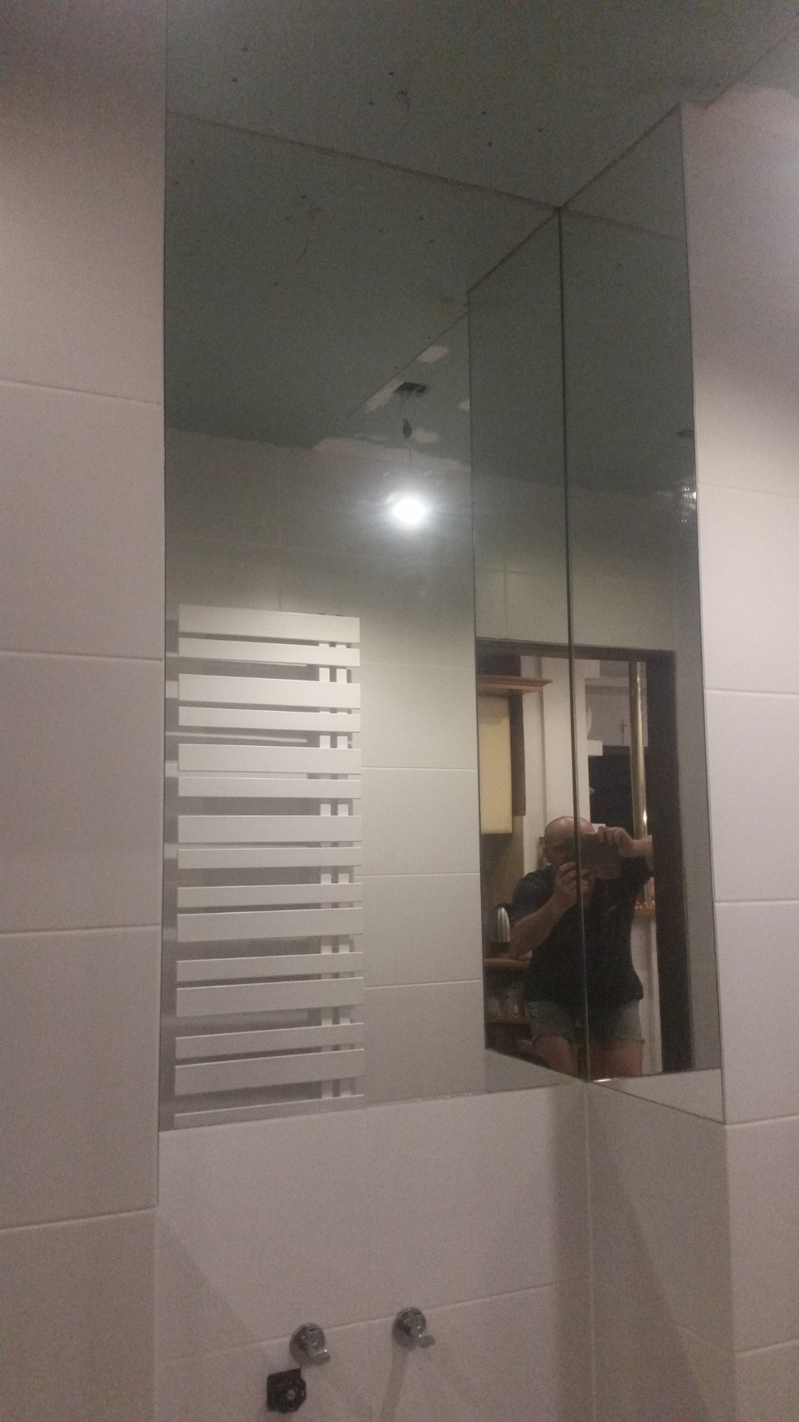 Biała łazienka z grzejnikiem drabinkowym odbijająca się w lustrze, widoczny fragment sufitu z niedokończonymi pracami