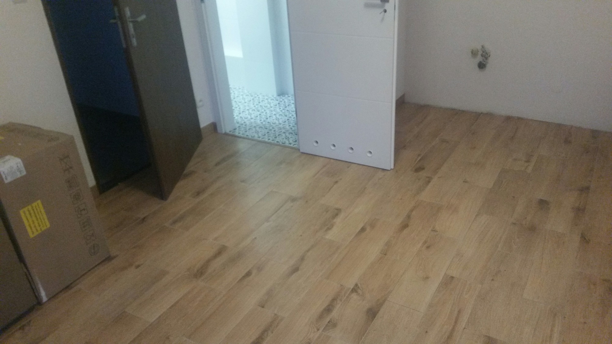 Ułożona podłoga z paneli imitujących drewno, widoczne otwarte drzwi do pomieszczenia z mozaikową podłogą, kartony w rogu pokoju i wystające ze ściany kable instalacji elektrycznej.