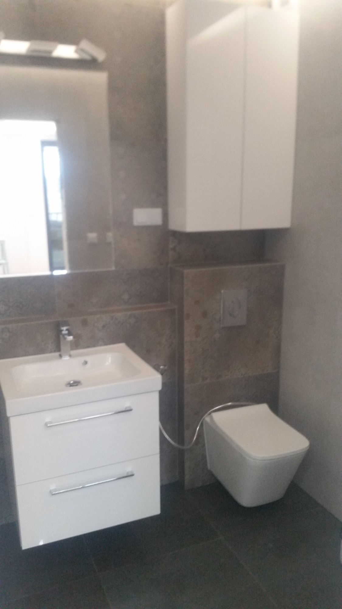 Nowoczesna łazienka z szarą płytką, podwieszaną toaletą i białą szafką z umywalką, minimalistyczny styl.