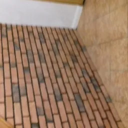 Ukośne ujęcie podłogi wyłożonej cegiełkami w odcieniach czerwieni i szarości, z fugami w kolorze czarnym; ściana wyłożona płytkami w odcieniach beżu; fragment drewnianej barierki w lewym dolnym rogu.