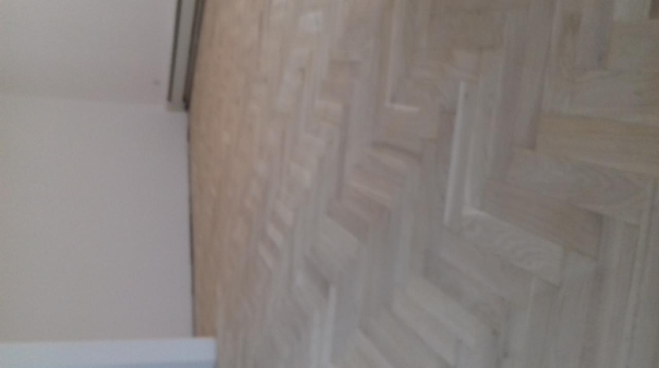 Jasna, drewniana podłoga ułożona we wzór jodełki francuskiej, widoczna z perspektywy ukośnej, z fragmentem białej ściany i listwy przypodłogowej w lewym górnym rogu kadru.