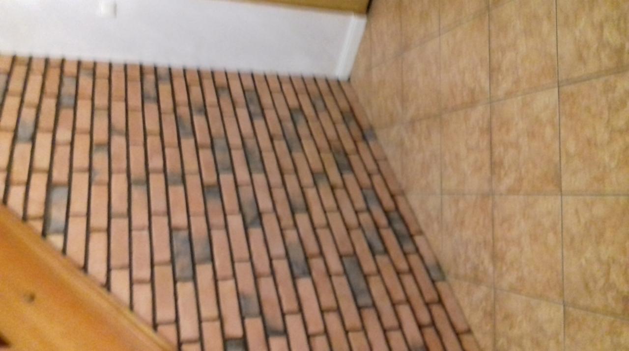 Ukośne ujęcie podłogi wyłożonej cegiełkami w odcieniach czerwieni i szarości, z fugami w kolorze czarnym; ściana wyłożona płytkami w odcieniach beżu; fragment drewnianej barierki w lewym dolnym rogu.