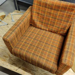 Fotel z pomarańczowo-brązową tapicerką w kratę, stojący na płycie OSB w warsztacie, widoczne elementy mocujące i gąbka w tle.
