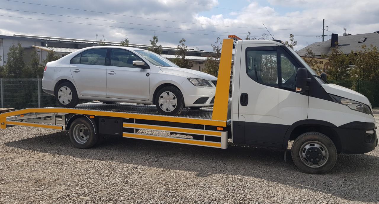 Srebrny samochód osobowy marki Skoda przewożony na platformie białej lawety z żółtymi elementami zabezpieczającymi, na żwirowym placu.