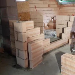 Pracownik budowlany w czapce z napisem 'PARTY HARD' niesie wiadro z zaprawą na budowie domu z bloczków ceramicznych Porotherm, widoczne ułożone bloczki i fragment ściany z drabinką.