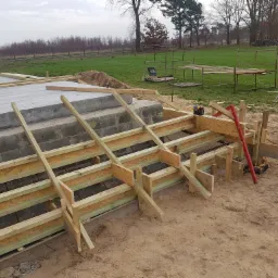 Betonowa podstawa w trakcie budowy z drewnianym szalunkiem, widoczne zbrojenie, poziomica i piła do drewna w tle, krajobraz z drzewami i trawnikiem.