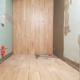 Montaż drewnopodobnych płytek na ścianie i podłodze w łazience, widoczne krzyżyki dystansowe, poziomica i instalacja wodna w ścianie, surowy stan wykończenia.