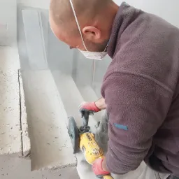 Pracownik w masce i rękawicach tnie szlifierką kątową płytę z betonu komórkowego, unoszący się pył, widoczne przygotowane płyty.