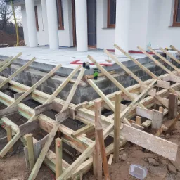 Szczegółowe ujęcie szalunku pod betonowy taras na etapie budowy, widoczna zbrojenie, poziomica laserowa i elementy drewniane, w tle biała elewacja domu z kolumnami.