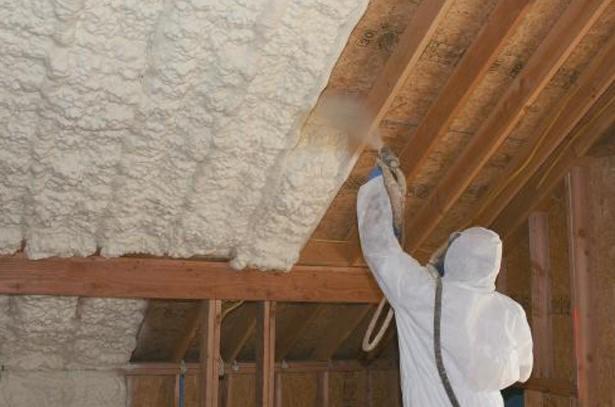 Aplikacja pianki PUR na poddaszu przez pracownika w kombinezonie ochronnym, widoczne drewniane elementy konstrukcji dachu i świeżo nałożona, nierówna warstwa izolacji.