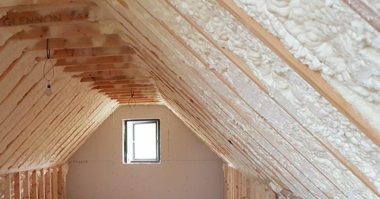 Poddasze domu w trakcie izolacji, widoczne drewniane krokwie pokryte warstwą białej pianki izolacyjnej, małe okno w szczycie, fragment instalacji elektrycznej z żarówką.
