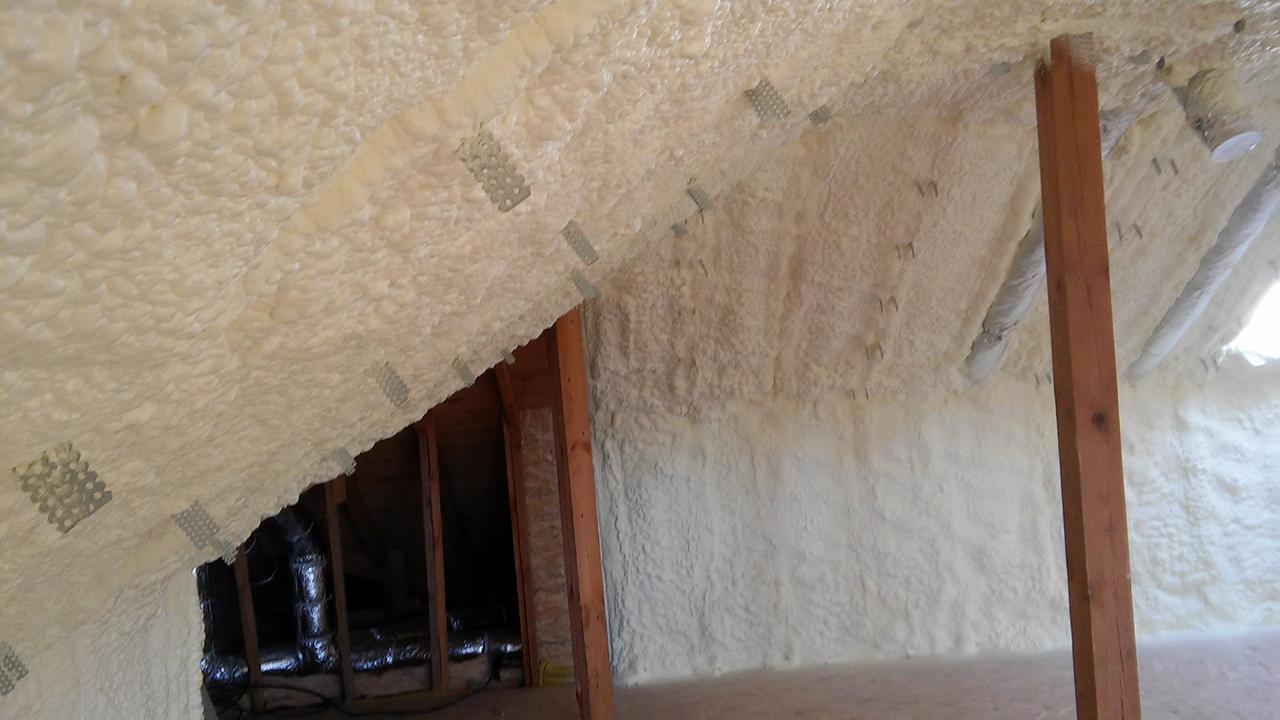 Poddasze po izolacji pianką: nierównomierna struktura piany pokrywa skośne ściany i sufit, widoczne drewniane elementy konstrukcyjne i metalowe łączniki, fragment instalacji wentylacyjnej.