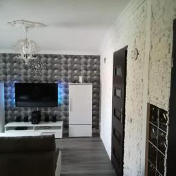 Usługi remontowo budowlane - Salon po metamorfozie: ściana z dekoracyjnej cegły w odcieniach bieli, tapeta 3D w geometryczny wzór, nowoczesne oświetlenie LED za telewizorem, białe meble i szara podłoga imitująca drewno.